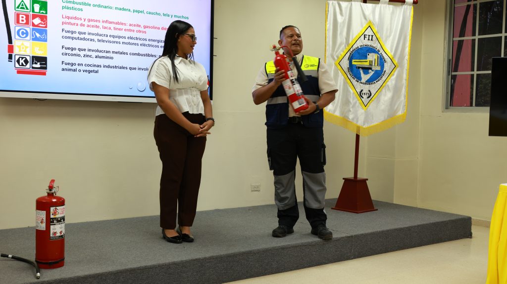 jornada de seguridad UTP 2026: especialistas de IISSO Management y Extintores Fire Rescue impartiendo el taller de prevención de incendios y uso de extintores, con la clasificación de tipos de fuego visible en pantalla y la bandera de la Facultad de Ingeniería Industrial de la UTP al fondo.
